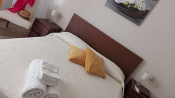 Una cama bien hecha con sábanas blancas y dos almohadas, una toalla doblada, una mesita de noche con una lámpara y un cuadro de orquídeas blancas colgado en la pared.