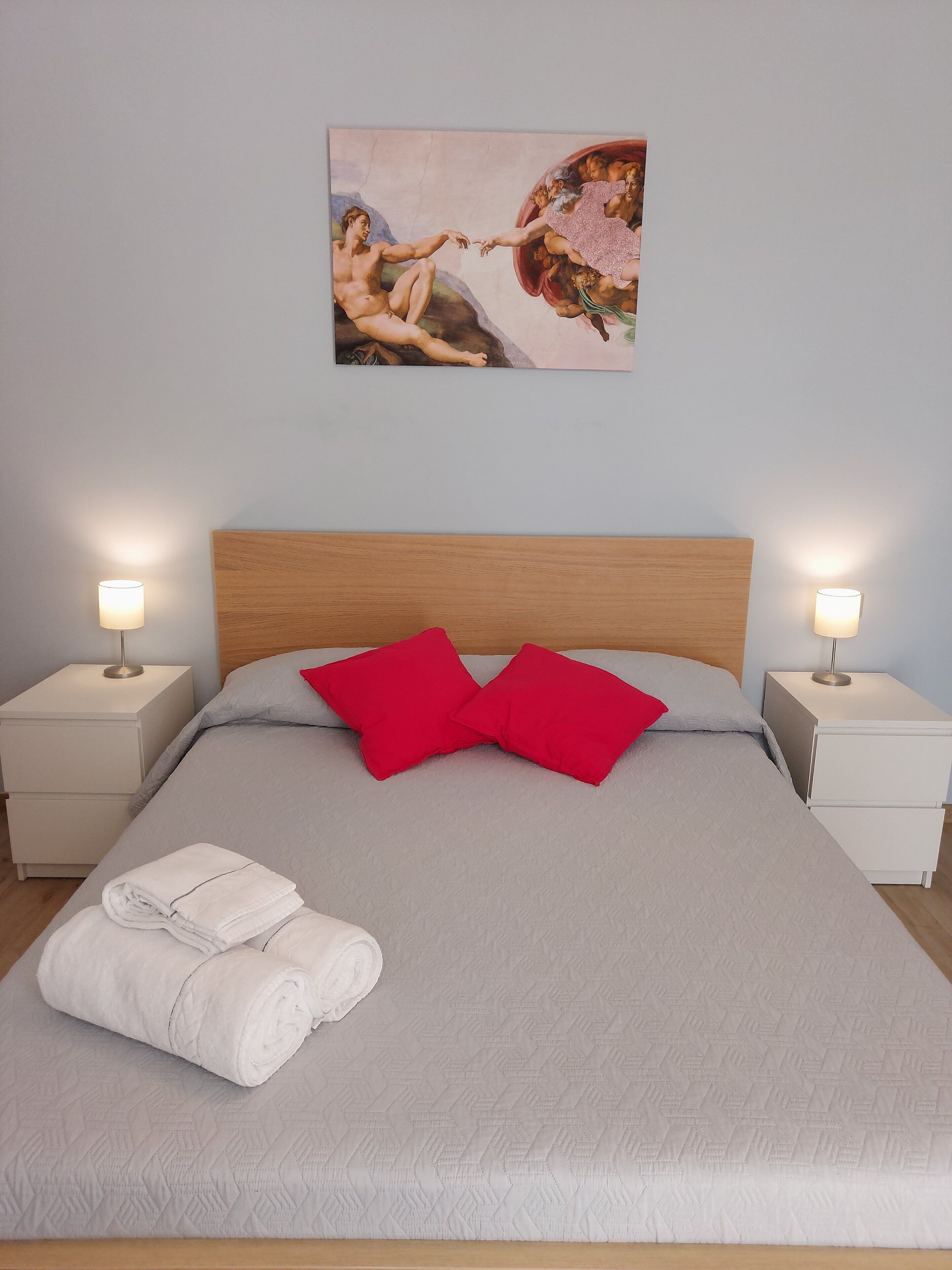 A neatly made bed with a grey quilt, two red pillows, and a white towel folded at the foot. There are two bedside tables with lamps, and a painting of cherubs on the wall.