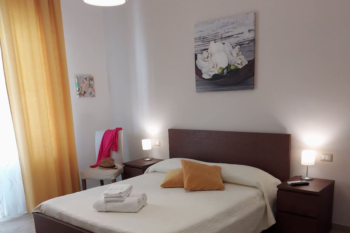 A neatly made bed with white and orange bedding, two bedside tables with lamps, a painting on the wall, and a wooden floor.