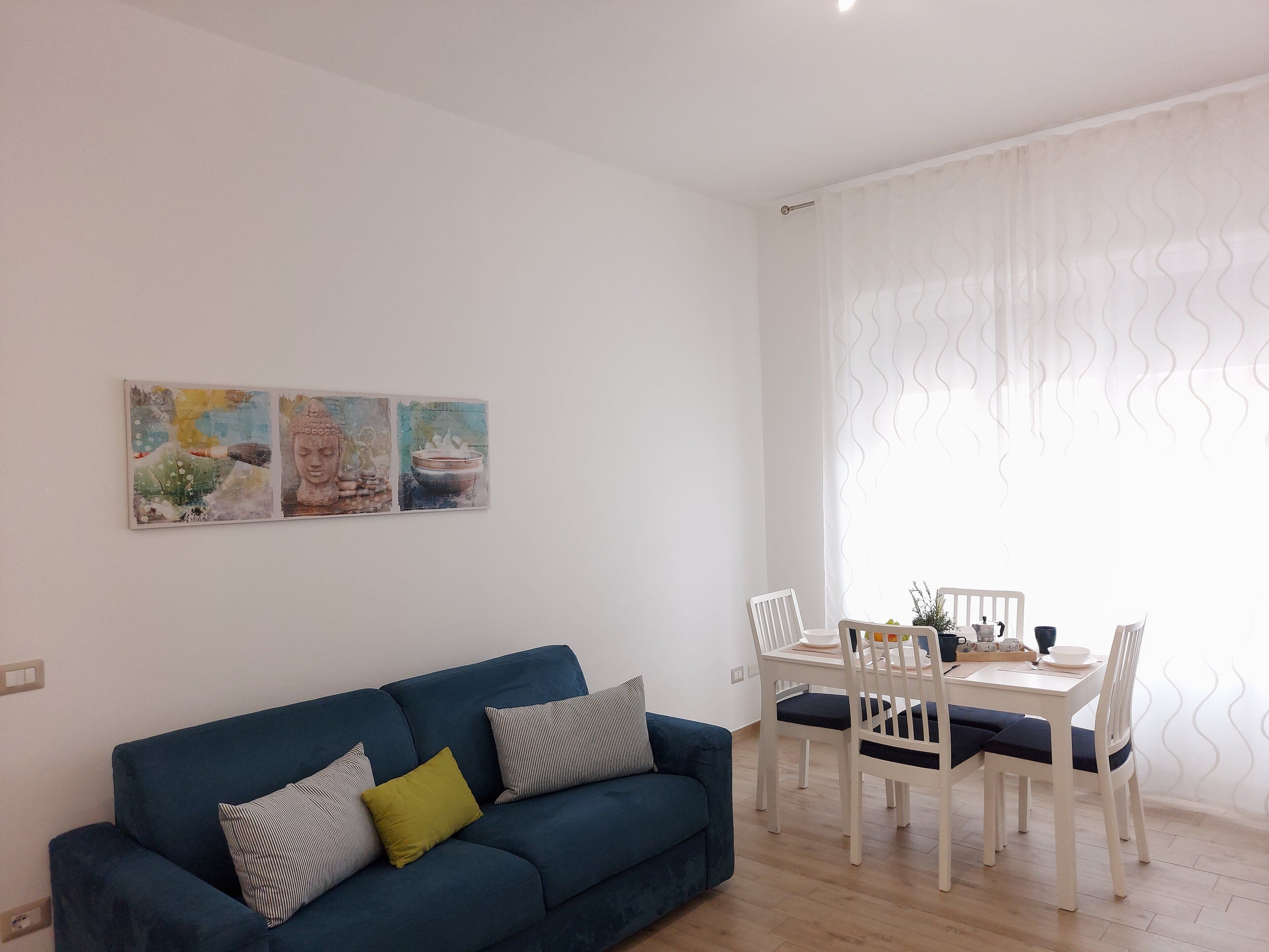 Een woonkamer met een blauwe bank, een witte eettafel en stoelen. Twee schilderijen hangen aan de muur.