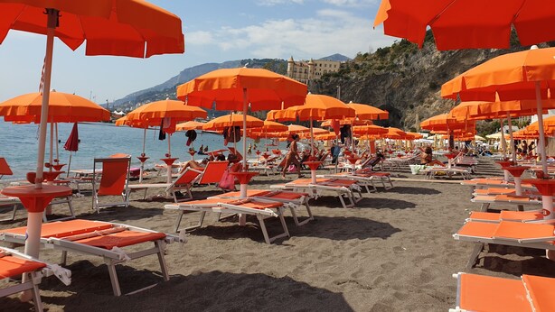 A beach with rows of orange sunbeds and umbrellas, a clear sky, and a mountainous backdrop. - Sapore di Mare Holidays (Maiori)