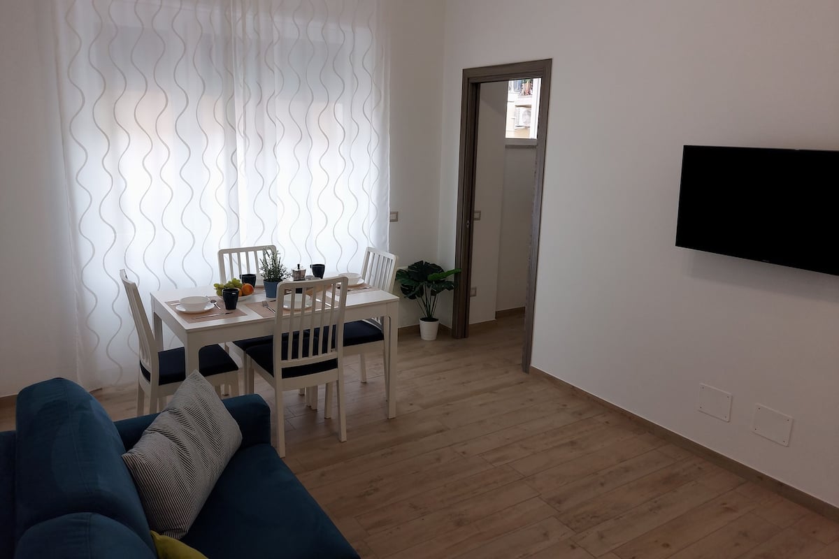 A living room with a blue sofa, a dining table with chairs, a potted plant, and a flat-screen TV mounted on the wall.