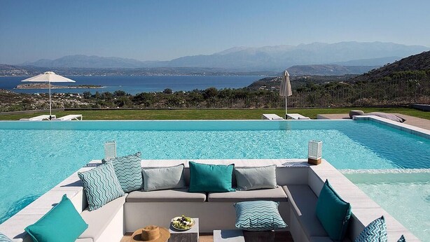 Zona junto a la piscina con un sofá y cojines, una mesa con frutero, y vista al mar y a las montañas.