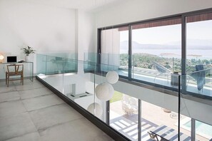 A modern interior with a glass balustrade, a desk with a laptop, and a view of a coastal landscape.