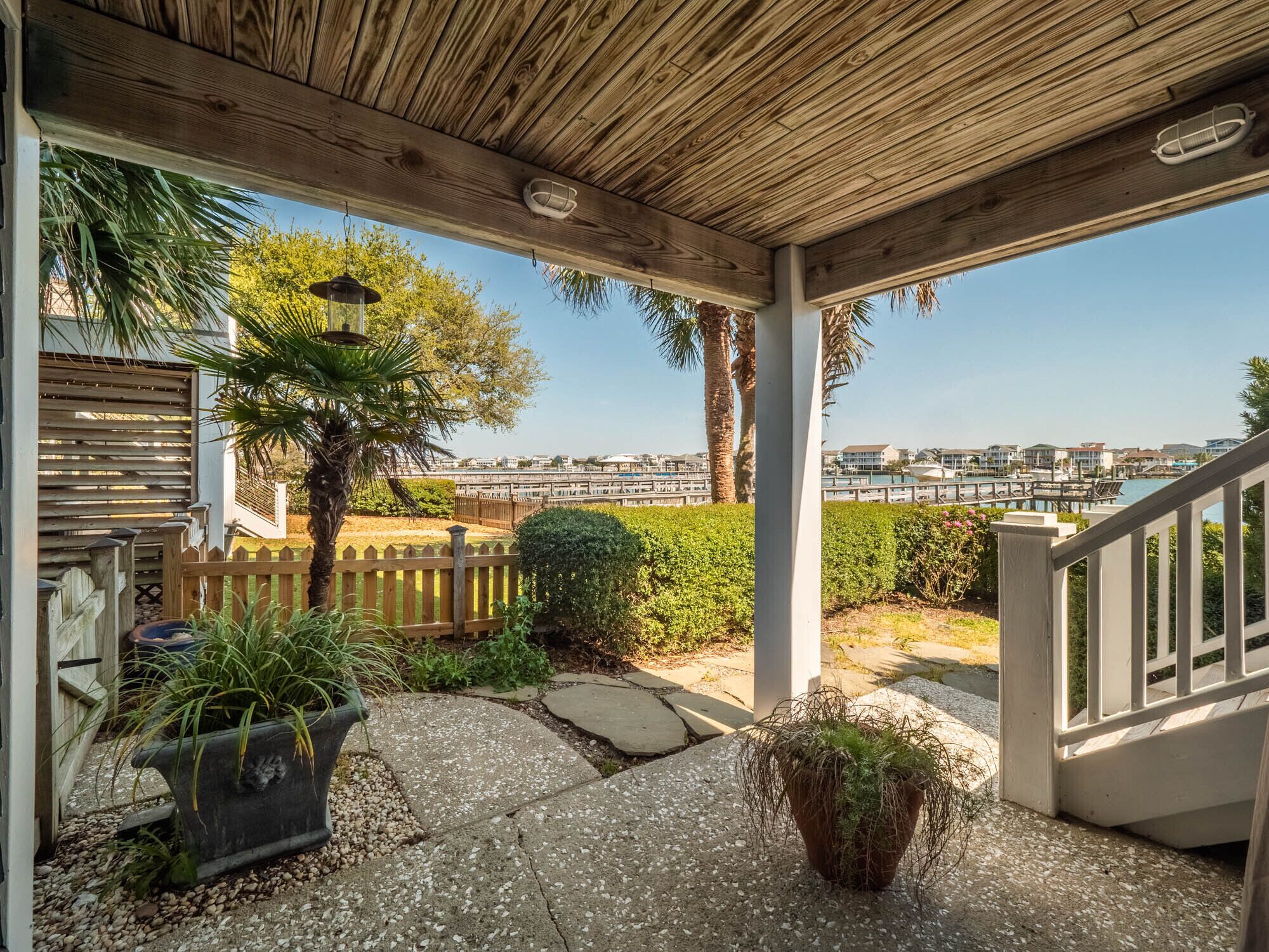 A covered outdoor area with a wooden ceiling, a wooden fence, and a view of a marina with boats and buildings in the background.