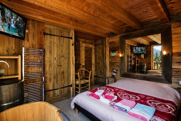 A wooden cabin bedroom with a bed, a wooden chair, a television, and a large mirror.