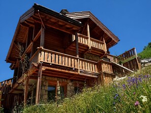 A wooden chalet with a balcony, stone foundation, and surrounded by wildflowers.
