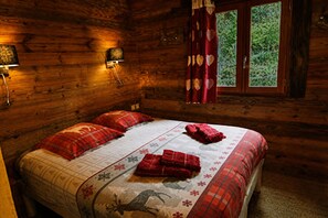 A wooden cabin bedroom with a bed, two pillows, a nightstand, a lamp, and a window with curtains.