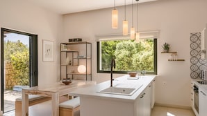 A modern kitchen with a central island, pendant lights, and a view of greenery outside. - Villa Rodia (Kefalonia)