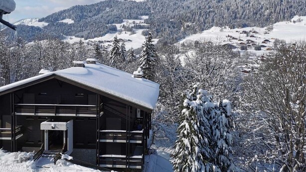 Une cabane dont le toit est recouvert de neige, avec un balcon et une vue sur un paysage montagneux enneigé.