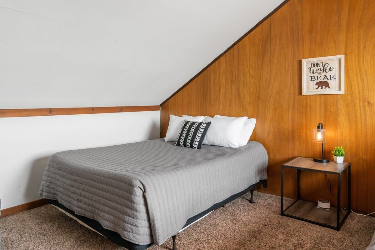 A bedroom with a sloped ceiling, a bed with a grey quilt, white pillows, a small bedside table with a lamp, and a framed quote on the wall.