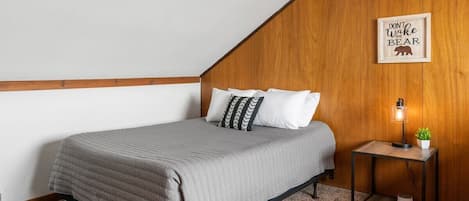 A bedroom with a sloped ceiling, a bed with a grey quilt, white pillows, a small bedside table with a lamp, and a framed quote on the wall.