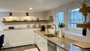 A modern kitchen with white cabinets, granite countertops, a built-in dishwasher, and a window with blinds.