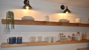A kitchen with wooden shelves holding plates, cups, and jars. There are hanging plants and pendant lights.