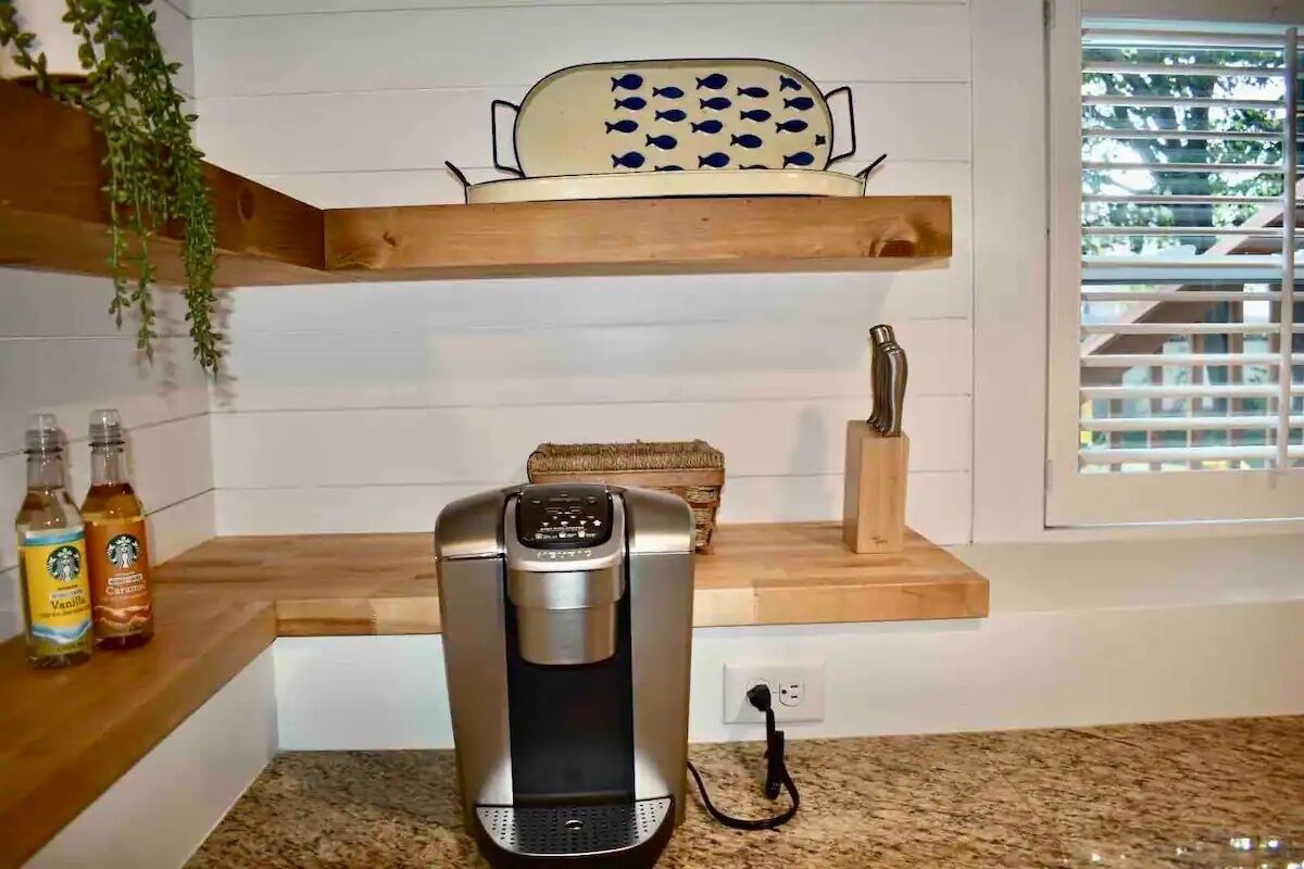 Une cuisine équipée d’une machine à café, d’une passoire ornée de motifs de poissons et de deux bouteilles de boissons posées sur le comptoir.