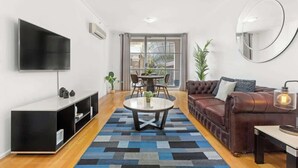 A modern living room with a flat-screen TV, a round coffee table, a leather sofa, and a dining area with a table and chairs.