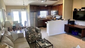 Un salon comprenant un canapé, un fauteuil et une table basse. Il y a une cuisine équipée d’un réfrigérateur, d’un four à micro-ondes et d’une cuisinière. On peut voir un ventilateur de plafond au-dessus de la cuisine.