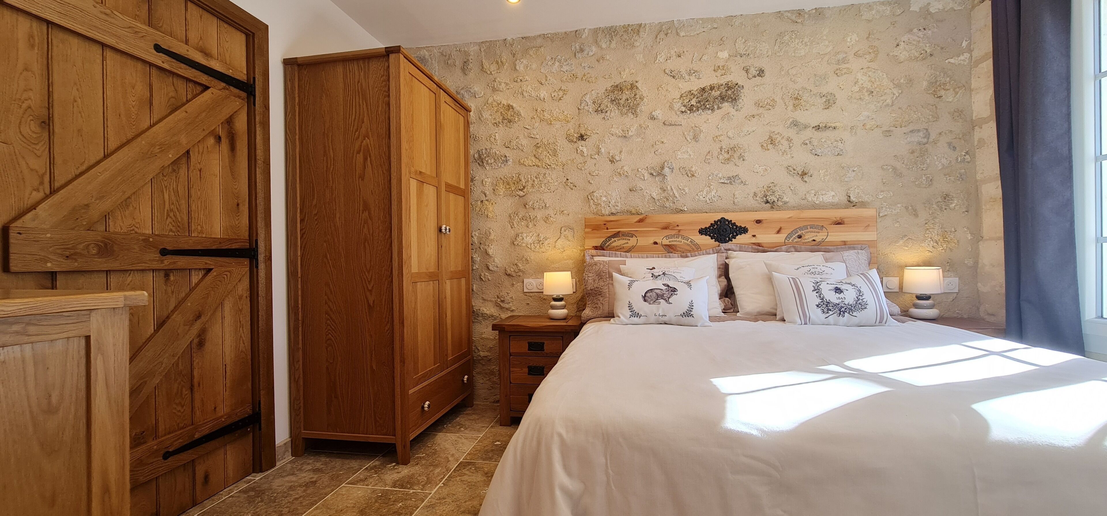 A bedroom with a large wooden wardrobe, a stone-textured wall, a wooden headboard, and a bed with multiple pillows.