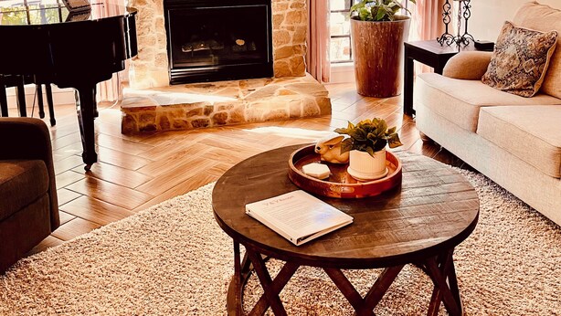 Una sala de estar acogedora con chimenea de piedra, un piano, una mesa de centro redonda con adornos y un sofá cómodo.