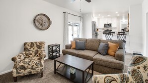 Un salon avec un canapé, un fauteuil et une table basse. On peut voir un ventilateur de plafond, et une cuisine se trouve à l’arrière-plan.