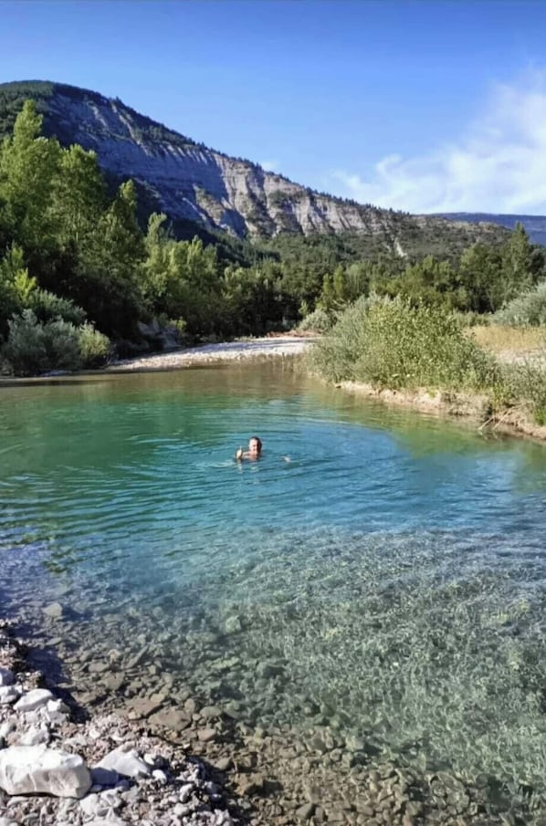 A clear river with a person swimming, surrounded by rocky terrain and greenery.