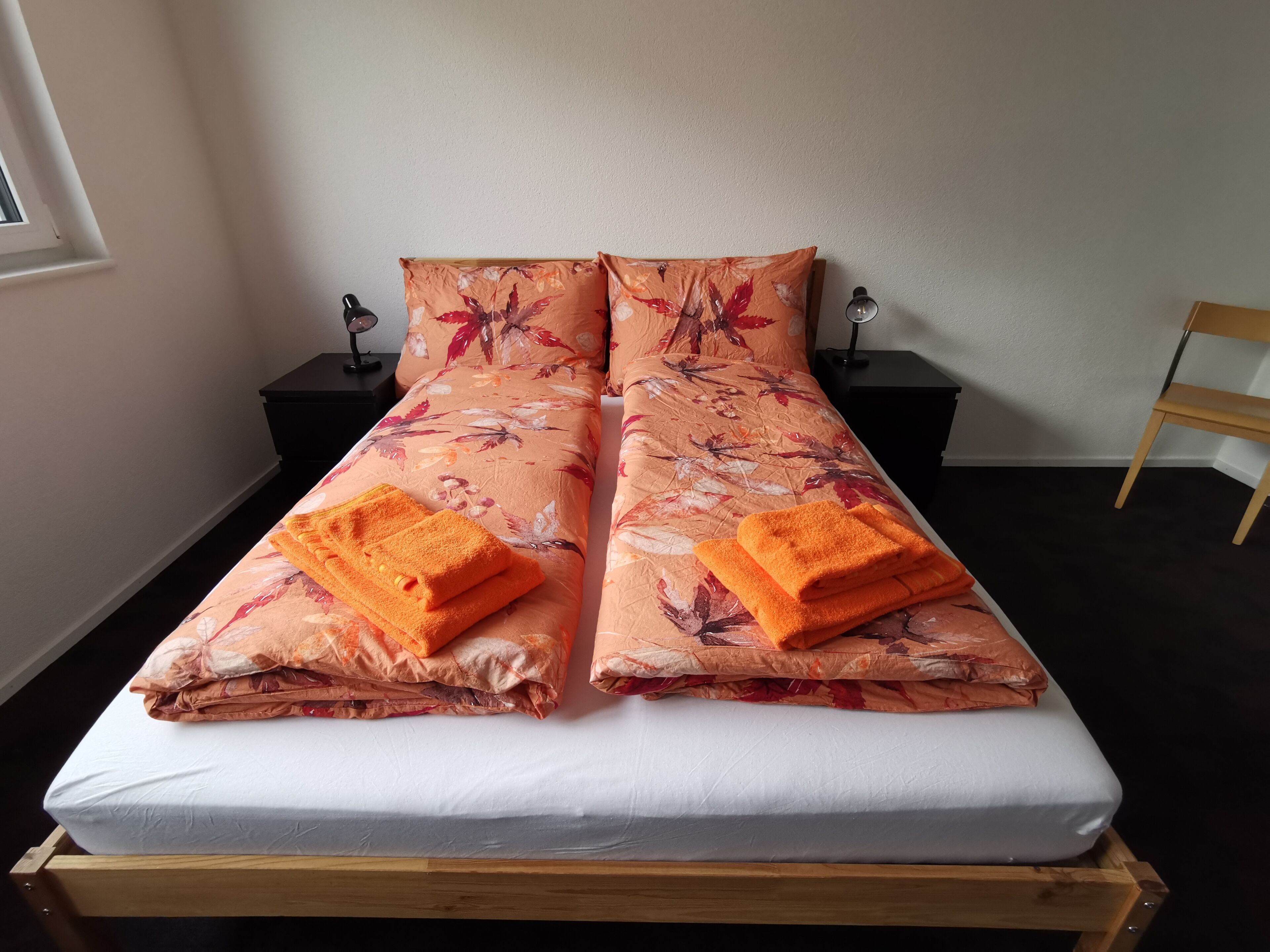 Ein Doppelbett mit floralem Überwurf und zwei orangefarbenen Handtüchern, ein Nachttisch aus Holz mit Lampe sowie ein Holzstuhl in einem Zimmer mit Fenster.