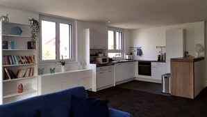 A modern kitchen with white cabinets, a microwave, and a dishwasher. There are bookshelves with books and decorative items. A blue sofa is in the foreground. - Ferienwohnung Hirschen (Gossau)