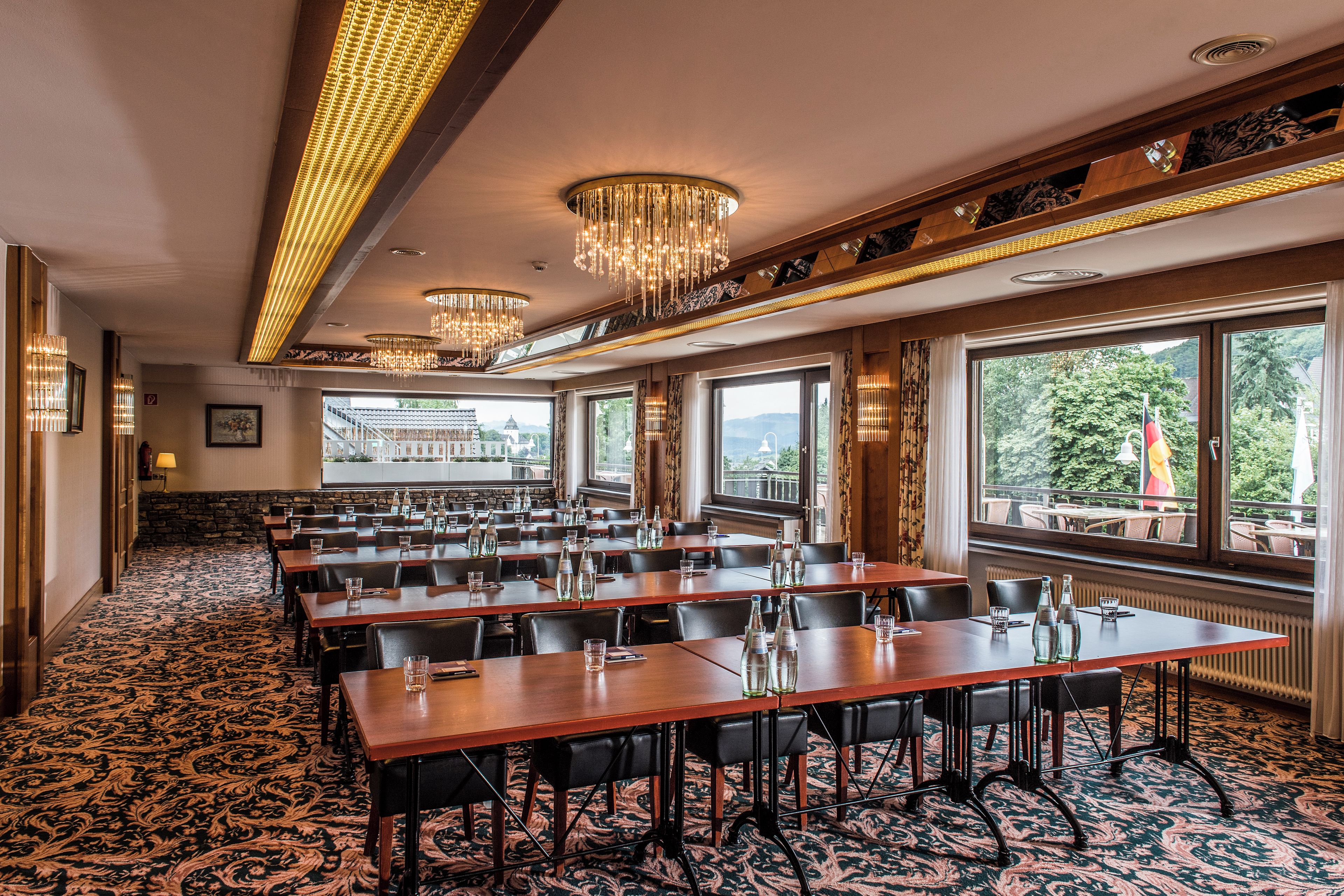 A conference room with multiple tables and chairs, large windows with a view of greenery, and ornate chandeliers.