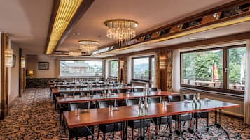 A conference room with multiple tables and chairs, large windows with a view of greenery, and ornate chandeliers.