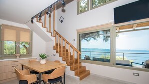 A modern kitchen with wooden flooring, a dining area with a wooden table and chairs, a staircase with wooden railings, and large windows offering a view of the sea. - Casa Spiaggia (Zakynthos - Limni Keriou)