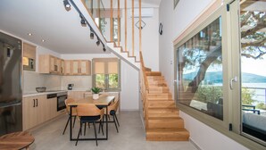 A modern kitchen with wooden cabinets, a dining area with a table and chairs, and a staircase leading to a balcony with a view of the outdoors. - Casa Spiaggia (Zakynthos - Limni Keriou)