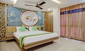 Un dormitorio con cama, cabecera de madera, ventilador de techo, pared con mural floral y cortina estampada.