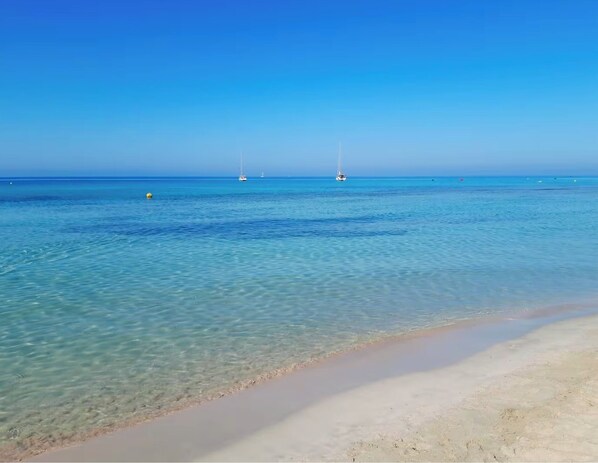 Une plage à l’eau claire, un rivage sablonneux et des voiliers à l’horizon.