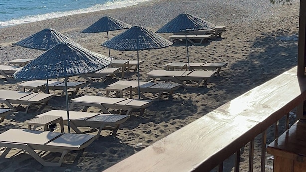 A beach with umbrellas and lounge chairs, a wooden deck, and a view of the sea and a mountain.