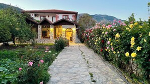 A stone pathway leading to a stone building with a red-tiled roof, surrounded by lush gardens and flowering plants.