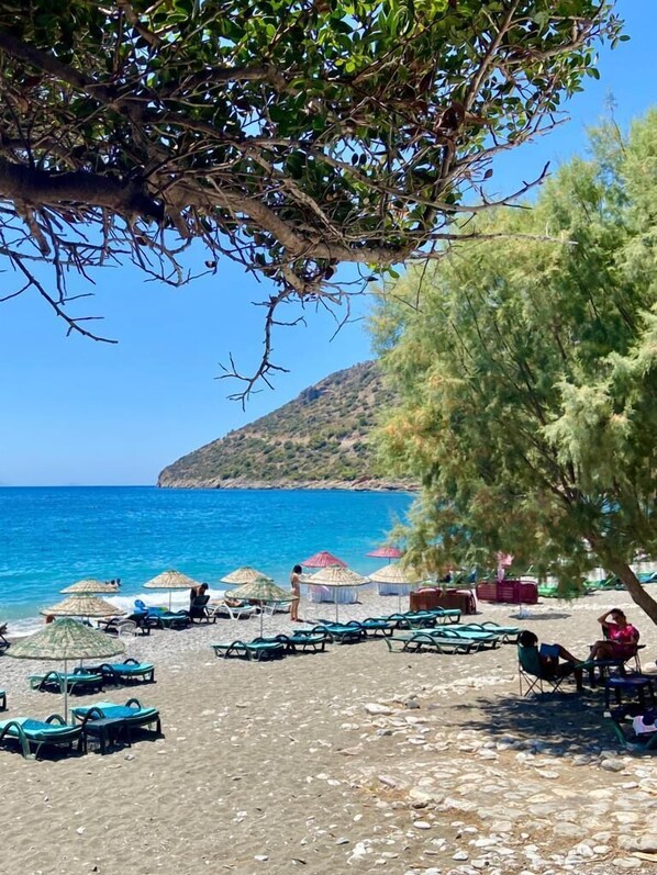 A beach with lounge chairs and umbrellas, a clear blue sea, and a hilly landscape in the background.