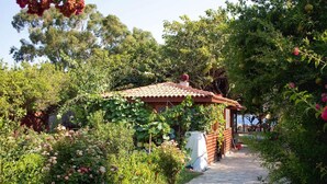 A rustic wooden cabin with a tiled roof, surrounded by lush greenery and flowering plants.
