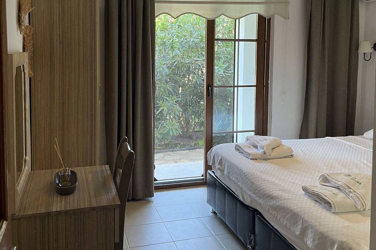 A bedroom with a bed, a wardrobe, a desk with a lamp, and a view of greenery outside.