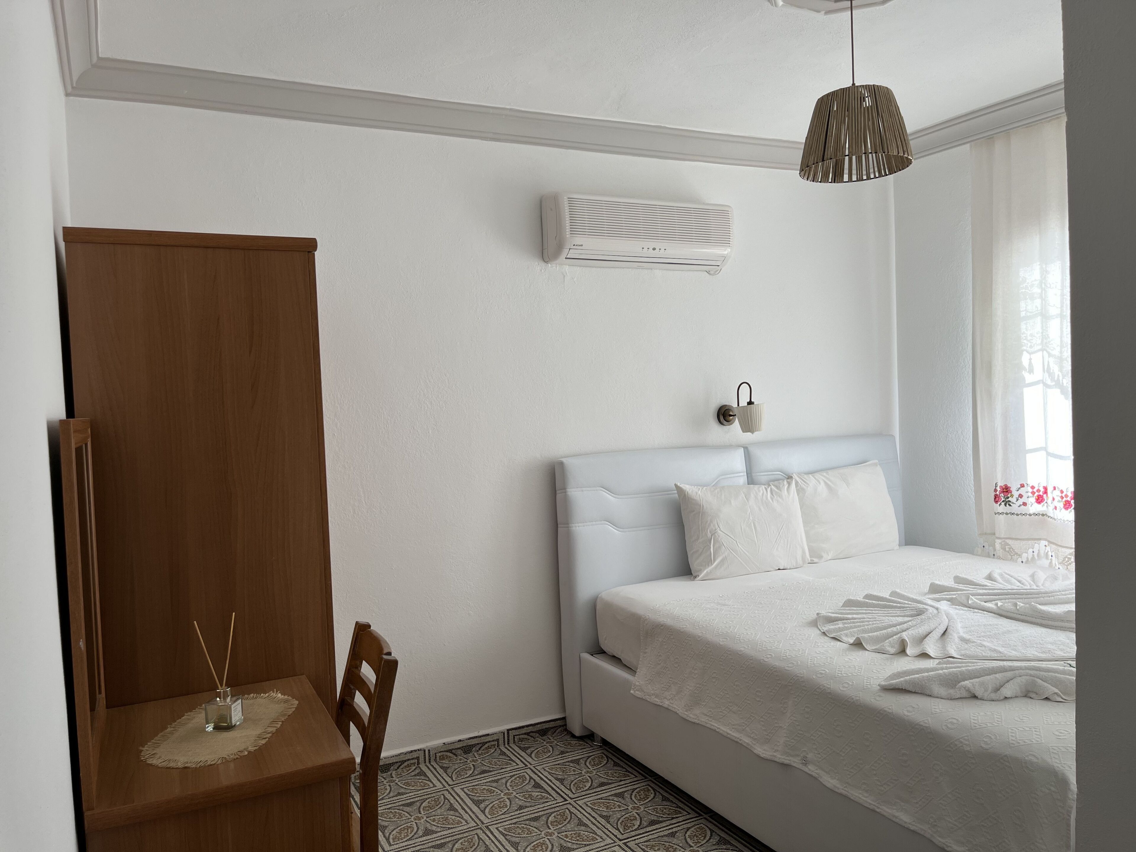 Un dormitorio con cama, mesita de noche, silla, aire acondicionado instalado en la pared y ventana con cortinas.