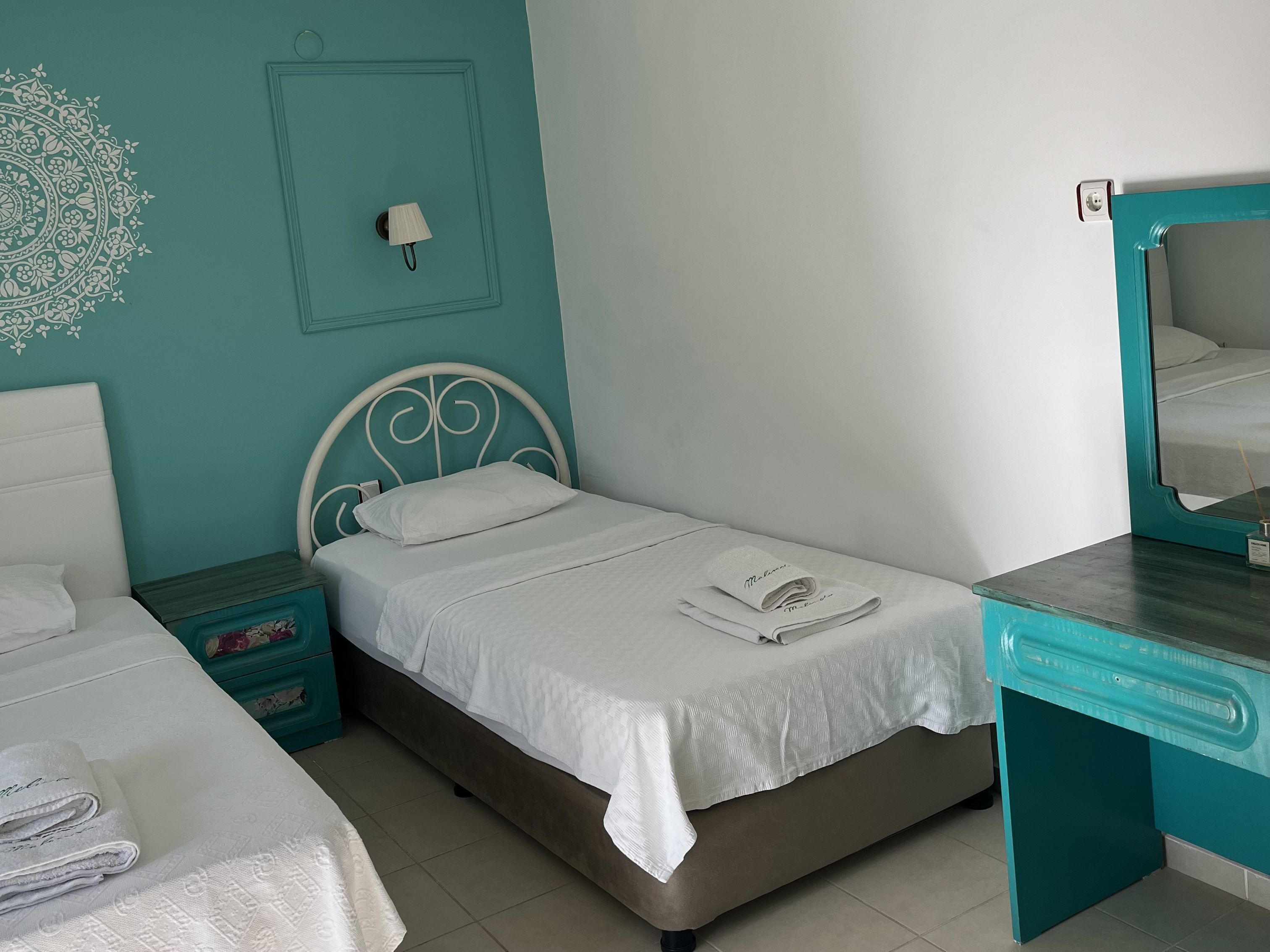 Una cama bien hecha con ropa blanca y una toalla doblada encima, una mesita de noche con una lámpara y un espejo en la pared contigua.