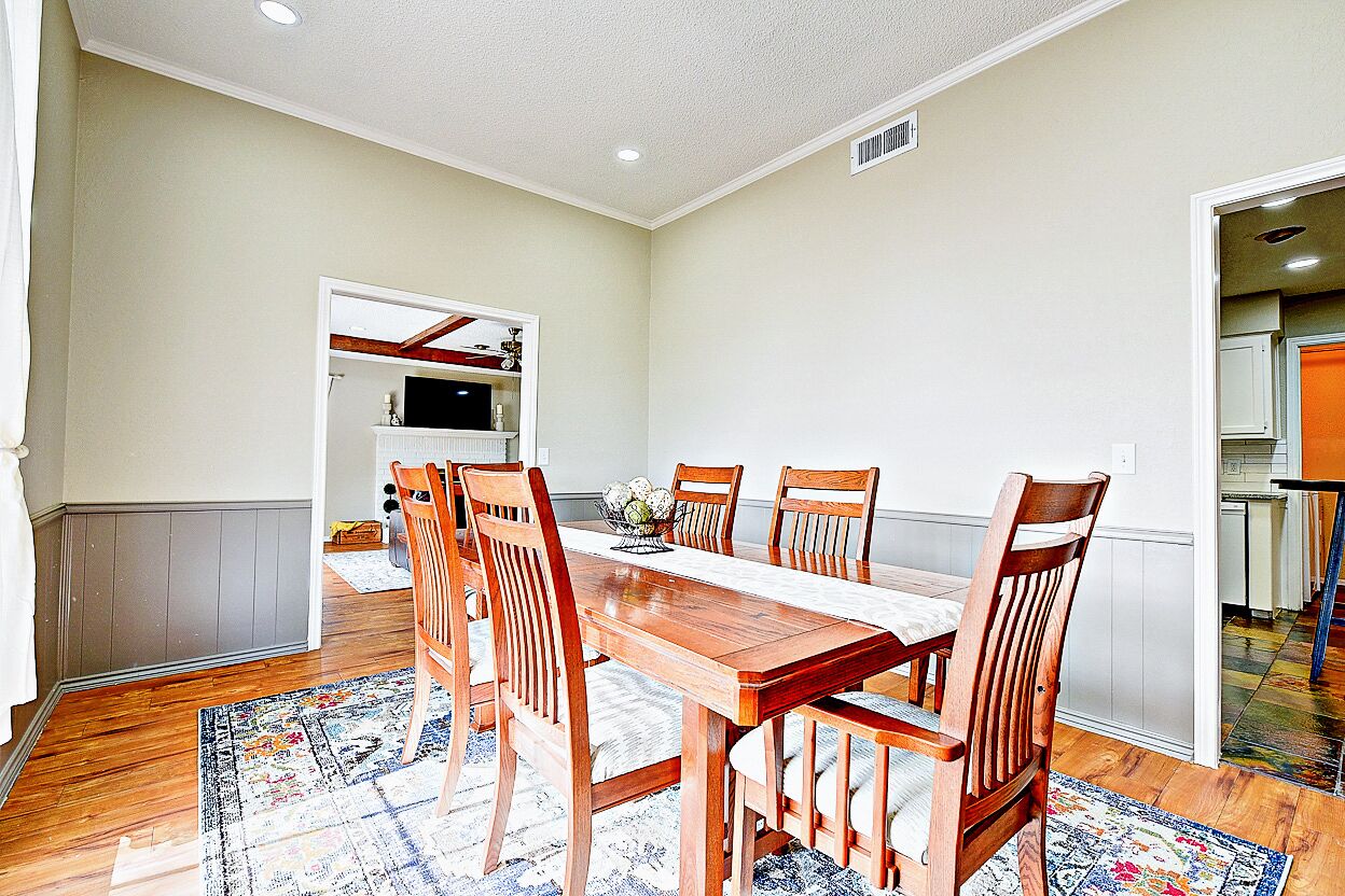 A dining area with a wooden table and chairs, a patterned rug, and a kitchen in the background.