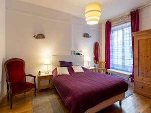 Quarto com uma cama grande, duas mesas de cabeceira com luminárias, uma cadeira de madeira, um guarda-roupa e uma janela com cortinas.