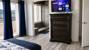 A hotel room with a bed, a dresser, a television displaying various channels, and a balcony with curtains. - Updated 1BR/1BA - "Hillside Getaway" (Lovenlund)