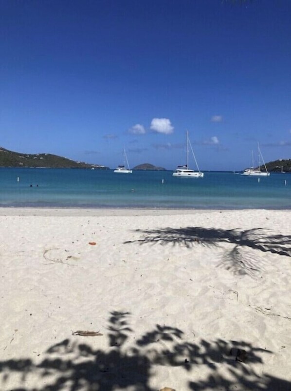 A beach with white sand and clear blue water. There are several sailboats anchored in the distance.