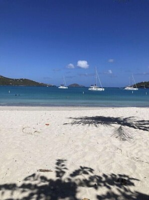 Une plage de sable blanc bordée d’une eau d’un bleu cristallin. On aperçoit plusieurs voiliers ancrés au loin.