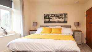A neatly made bed with white and yellow bedding, two bedside tables with lamps, a window with a view, and a painting on the wall. - Yippee Cottage (Keighley)