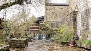 A stone-paved courtyard with a table and chairs, surrounded by stone walls and vegetation. - Yippee Cottage (Keighley)