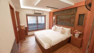 A bedroom with a large bed, wooden flooring, a window with blinds, and a wall-mounted air conditioner.