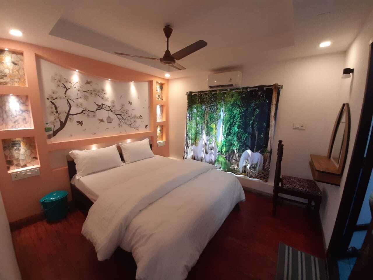 A bedroom with a bed, a ceiling fan, a wall with a tree mural, a window with a forest scene, and a small table with a chair.