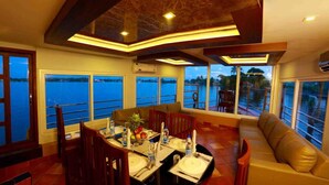 A dining area with a table set for six, large windows offering a view of the water, and a seating area with a sofa. - Double Decker Houseboat (Ambalapuzha)